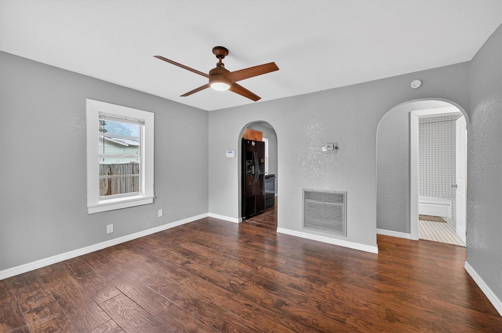1908-3 - Graystone Investment Group Living room with gray walls, dark wood floor, and a wooden ceiling fan; window shows an outdoor yard and a doorway to the kitchen and bath via arched openings.