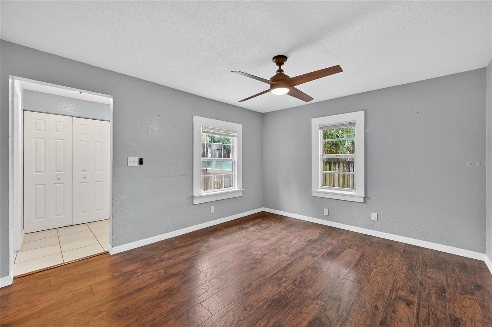 1908-4 - Graystone Investment Group Empty living room with gray walls, two windows, and dark wood floors, light fixture ceiling fan center stage.