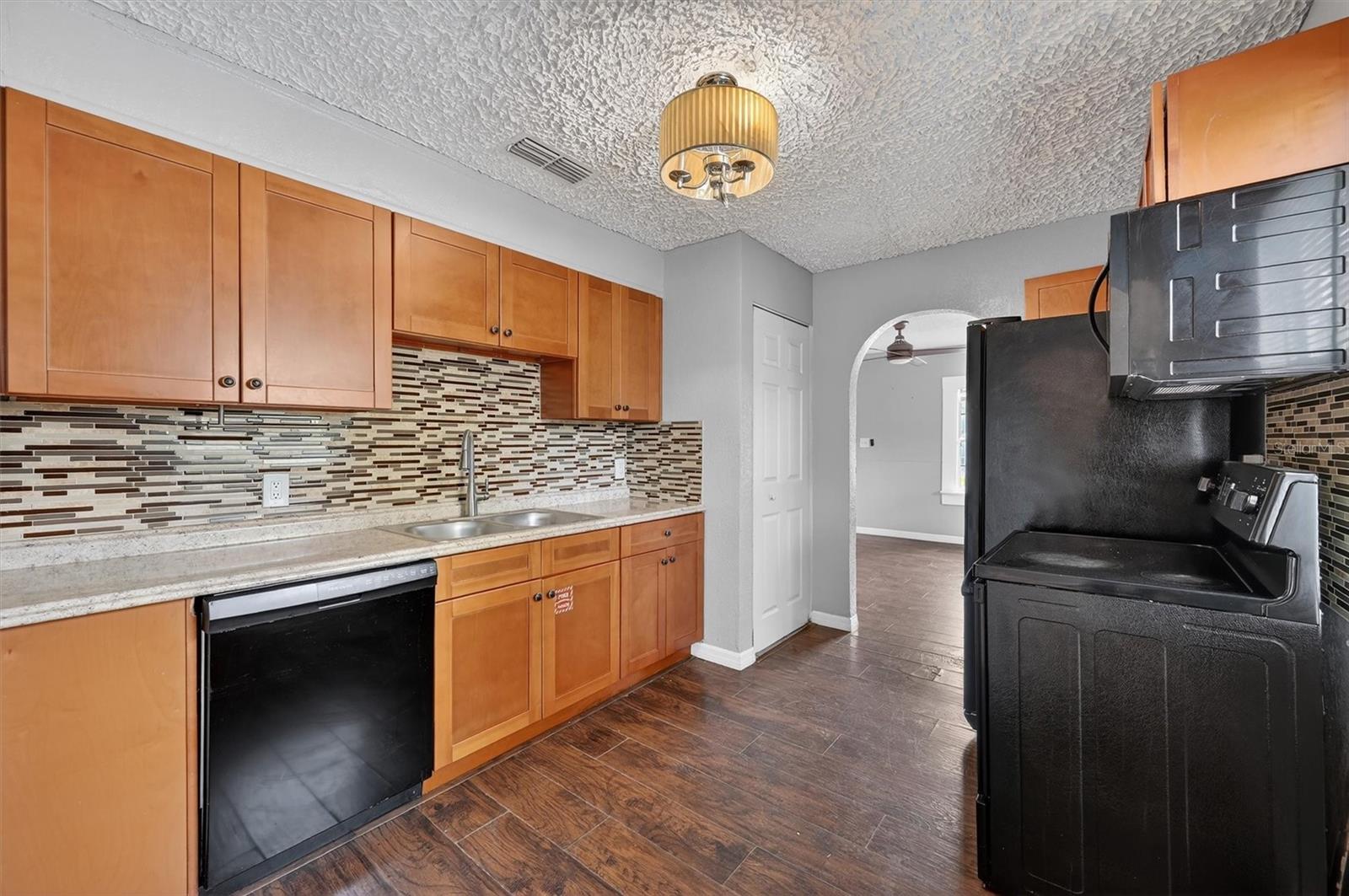 1908-7 - Graystone Investment Group Modern kitchen with wooden cabinets, mosaic tile backsplash, double sink, and black appliances (dishwasher and range). Recessed ceiling light above center.