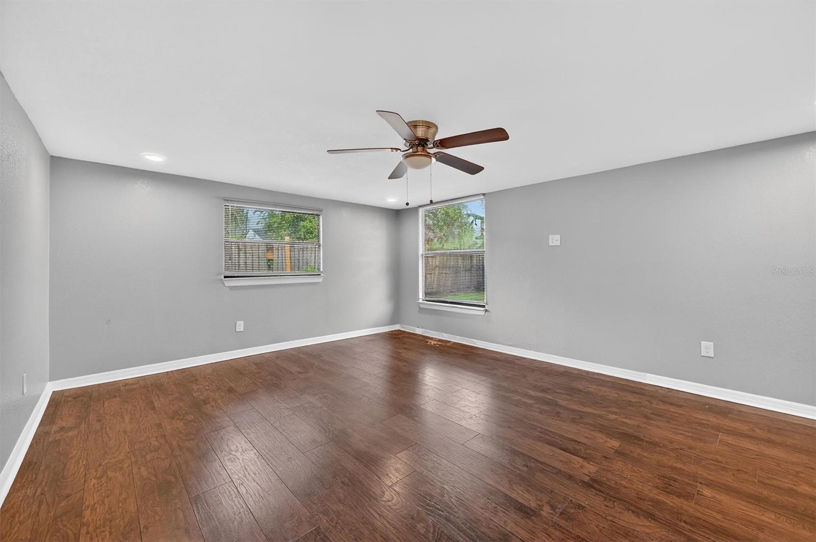 1908-9 - Graystone Investment Group Bright empty basement room with gray walls, dark wood floors, two windows, and a ceiling fan.