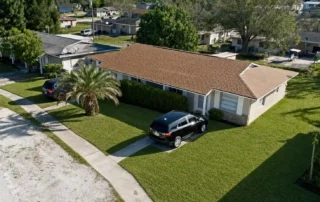 Aerial view of a single-story house with a brown shingled roof, two cars in the driveway, and a palm tree in a green front yard.
