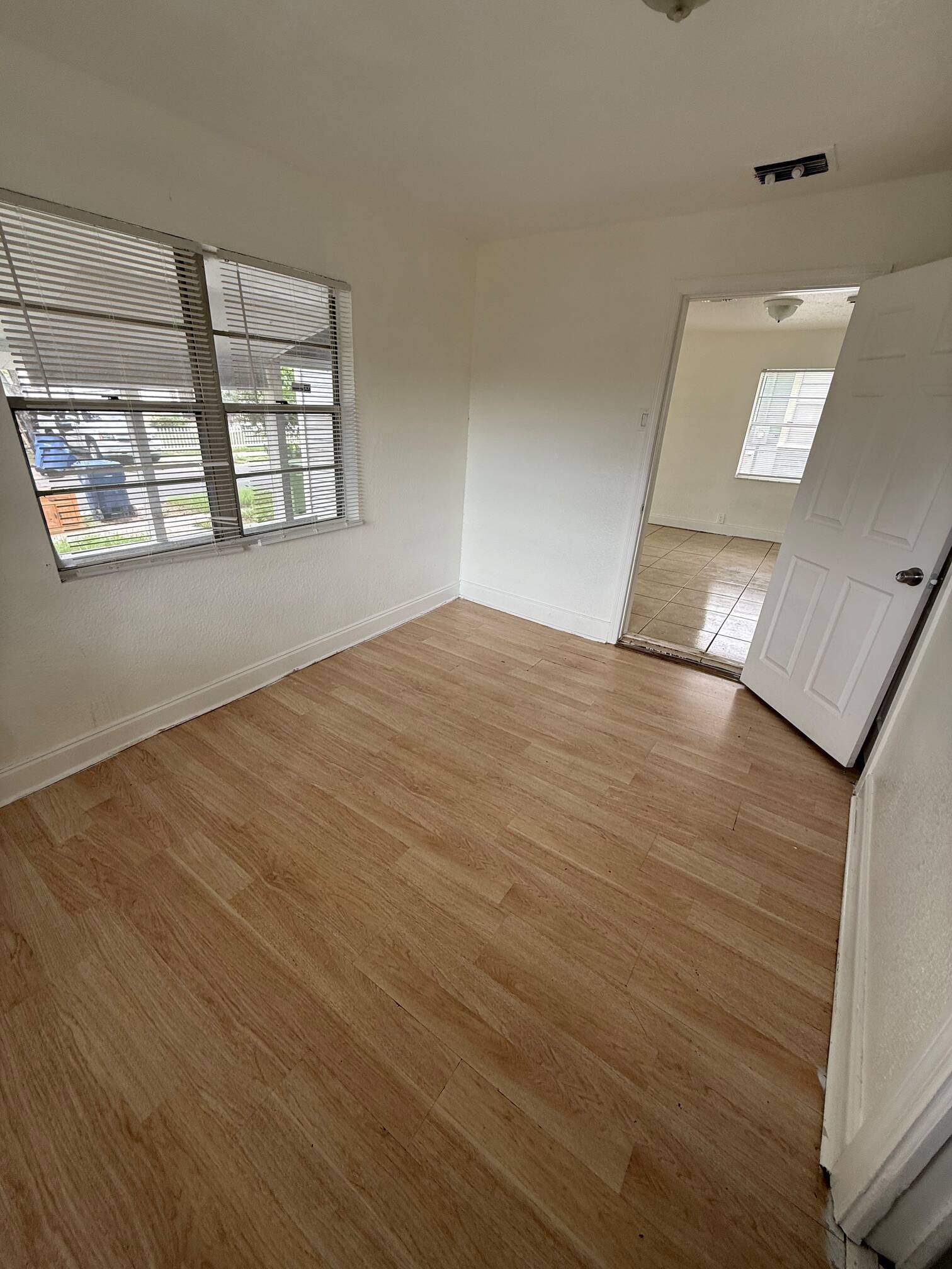 WhatsApp Image 2026-07-23 at 61537 PM - Graystone Investment Group Small empty room with white walls, light wood floor, window with blinds, and an open door to a tiled adjacent room.