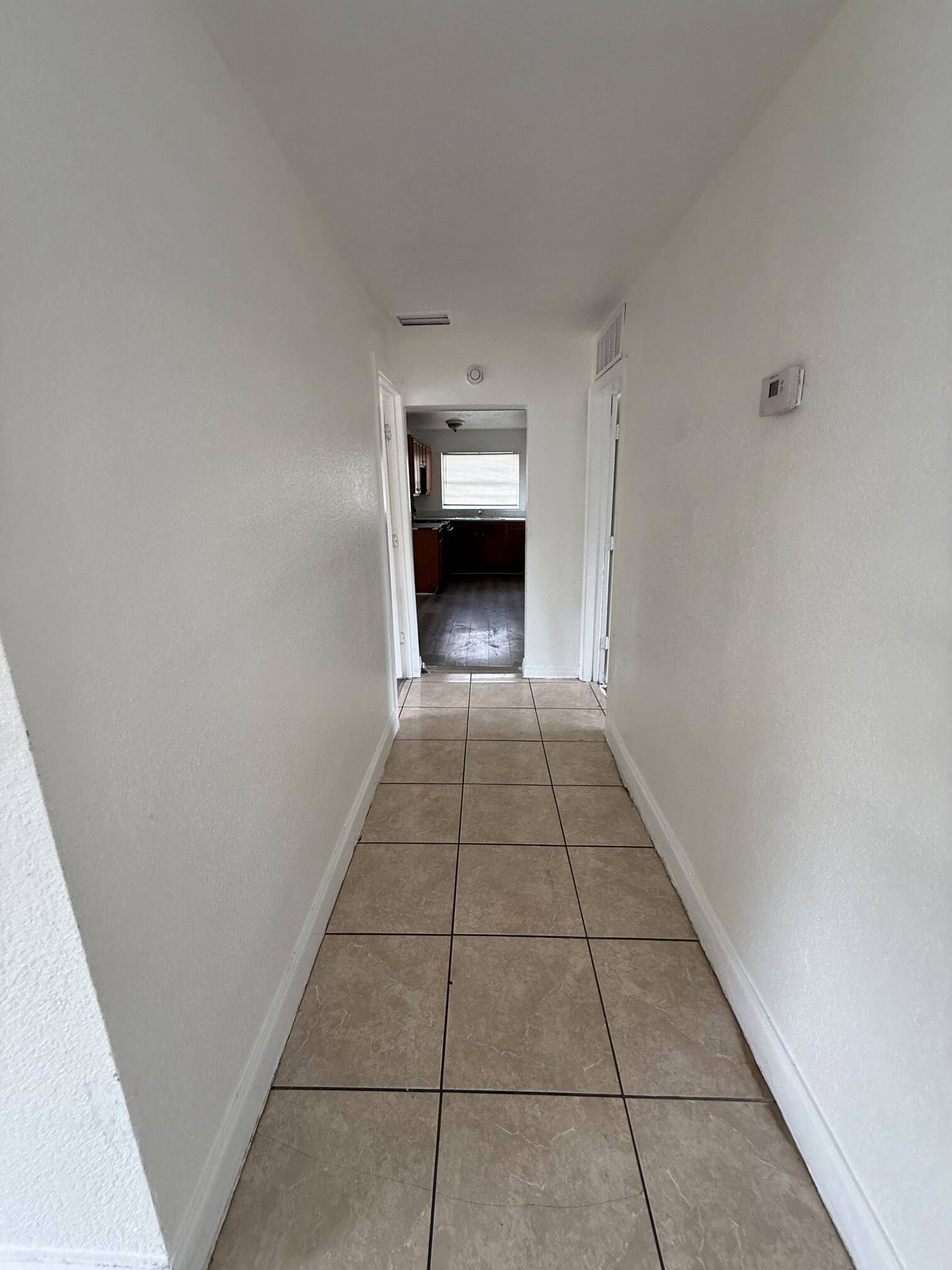 WhatsApp Image 2026-07-23 at 61539 PM - Graystone Investment Group Narrow hallway with beige tiled floor and white walls, extending to a kitchen at the far end with a window.
