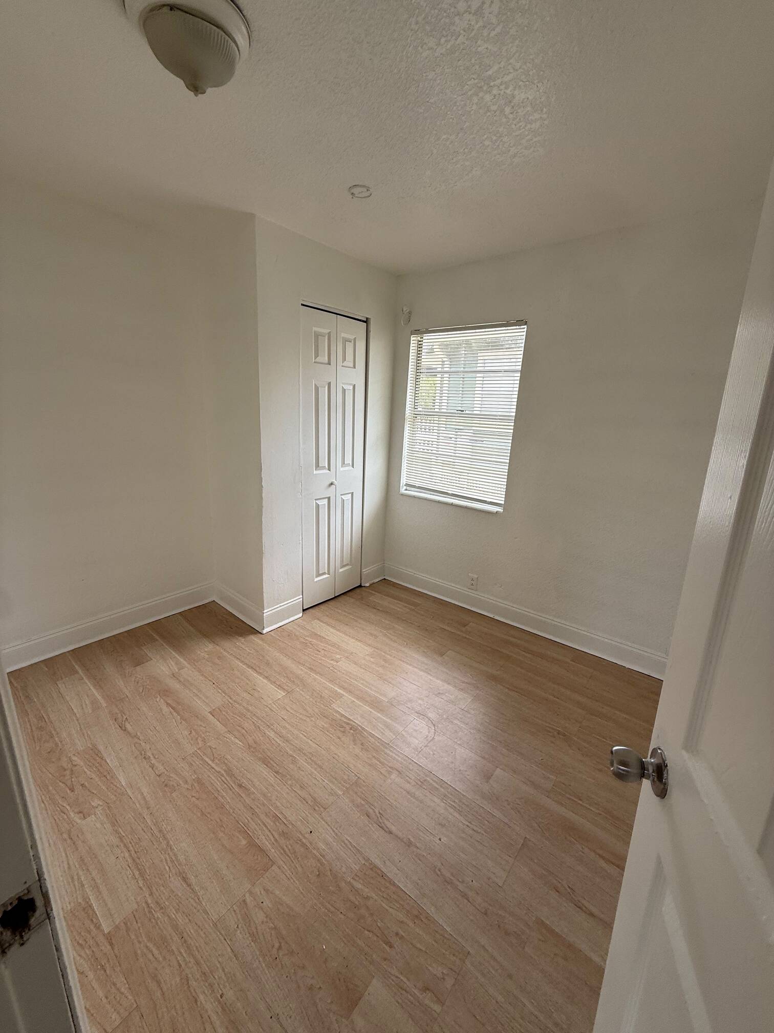 WhatsApp Image 2026-07-23 at 61541 PM - Graystone Investment Group Empty small room with wood-like flooring, white walls, window with blinds, and a closed closet door on the left.