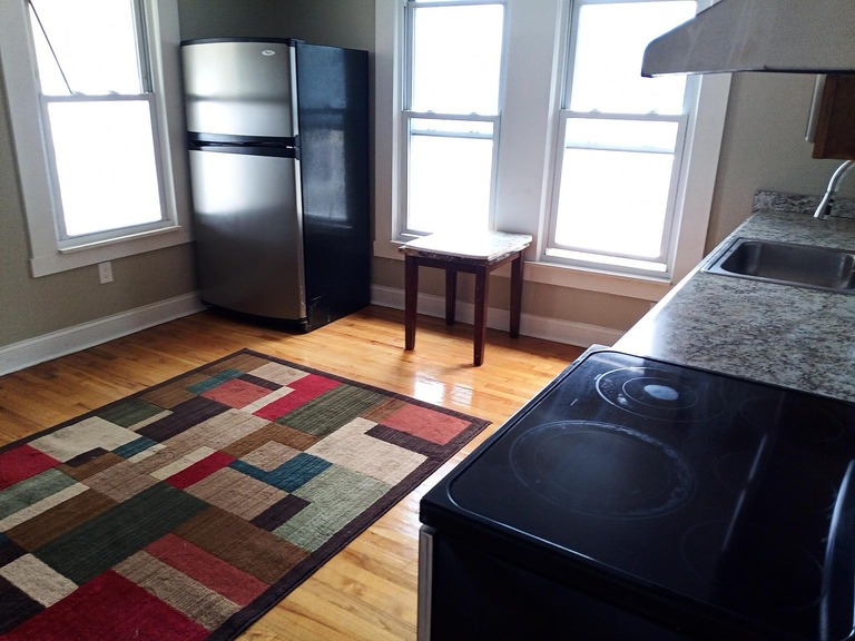 imgi_670_6402dc6bda41f160879627754d1b0291-uncropped_scaled_within_1536_1152 - Graystone Investment Group Cozy kitchen with a stainless steel refrigerator, wooden floor, and a colorful geometric rug by a small table near three windows