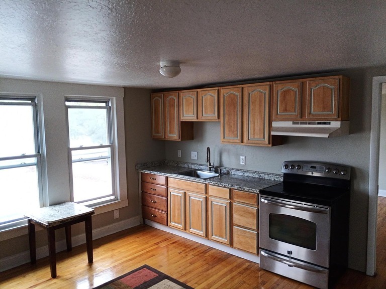 imgi_678_1f49e1fd6ad311f11d21ef7fba7cbf9f-uncropped_scaled_within_1536_1152 - Graystone Investment Group Kitchen with oak cabinets, granite counters, stainless steel stove, and a small table by two sash windows on a hardwood floor.