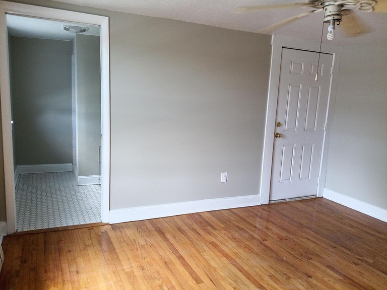 imgi_710_0a3eed9914350e954b3857b23fe5d206-uncropped_scaled_within_1536_1152 - Graystone Investment Group Empty living room with honey‑colored wood floor, light gray walls, white baseboards, and a white door; doorway to a small tiled area on the left.