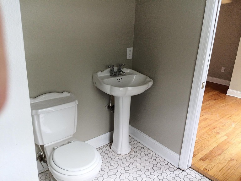 imgi_718_55713f6df9ca975201eddfde8143660a-uncropped_scaled_within_1536_1152 - Graystone Investment Group A small bathroom with a white toilet and a pedestal sink against light gray walls and white hex tile floor edge-by-side, near a doorway to a wooden floor room.