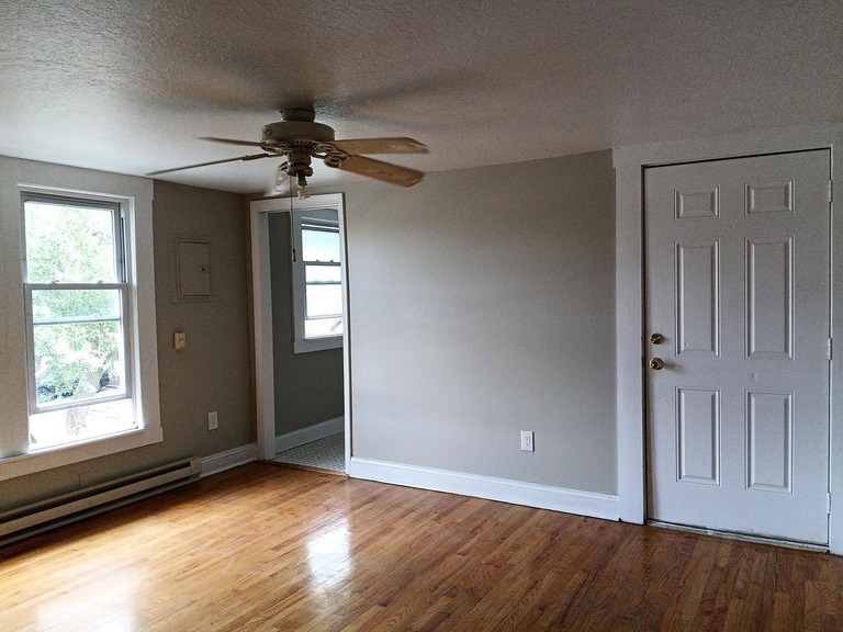 imgi_726_cd54d73cac2d661a89cf2398a898b7d9-uncropped_scaled_within_1536_1152 - Graystone Investment Group Living room with light hardwood floors, a white ceiling fan, two front windows, baseboard heater, and a white entry door on the right.