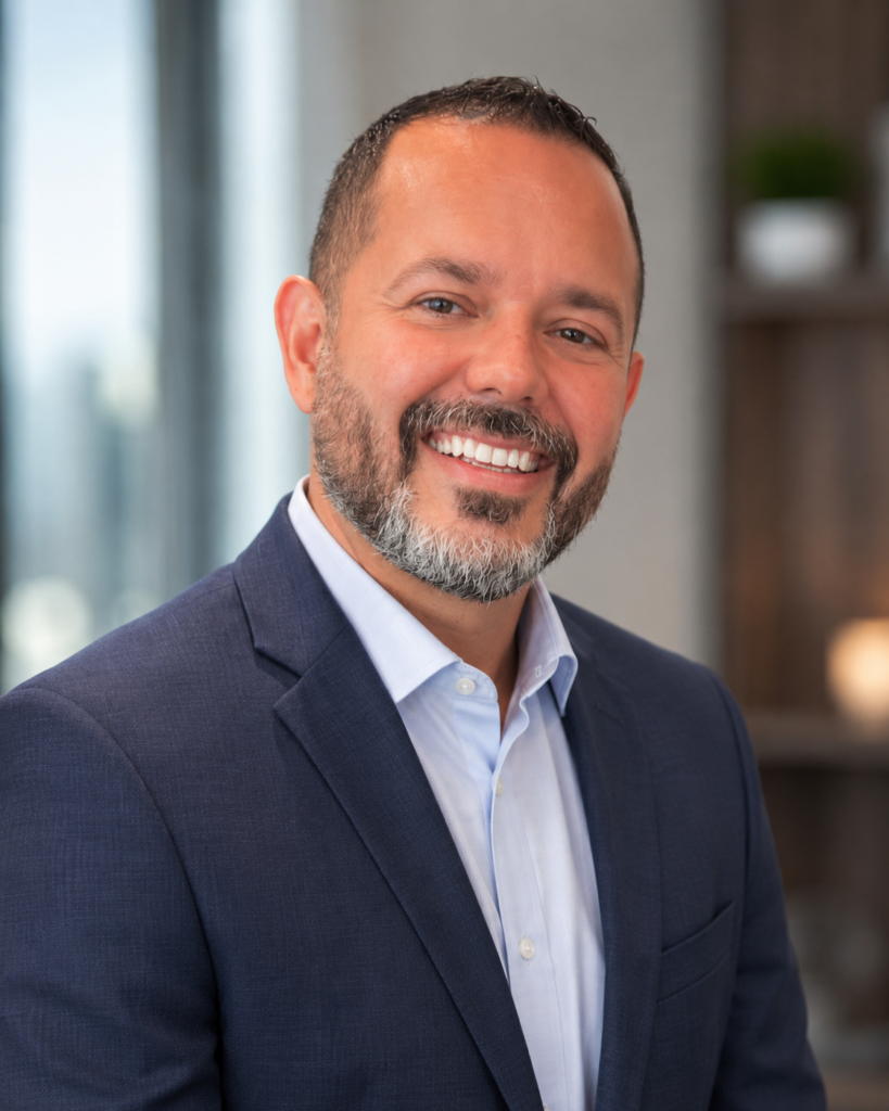 Smiling man in a navy blazer and light blue shirt posing for a professional headshot in an office setting.