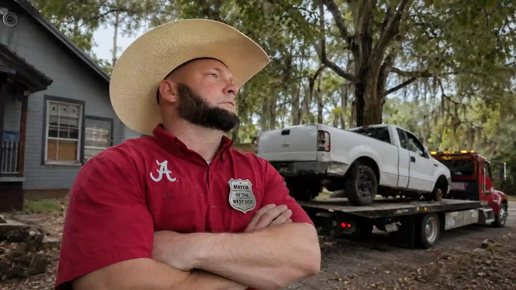 Bearded man in a wide-brimmed straw hat and red shirt with a badge reading 'Mayor of the West Side' stands with arms crossed beside a tow truck carrying a white pickup in a tree-filled yard.